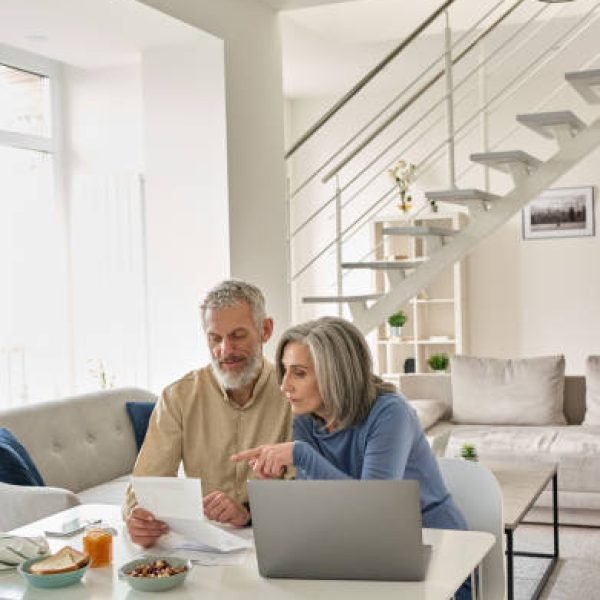 Senior mature couple check bank loan documents using laptop at home. Middle aged old family read paper bills payments, calculating pension or taxes, planning retirement finances, household expenses.